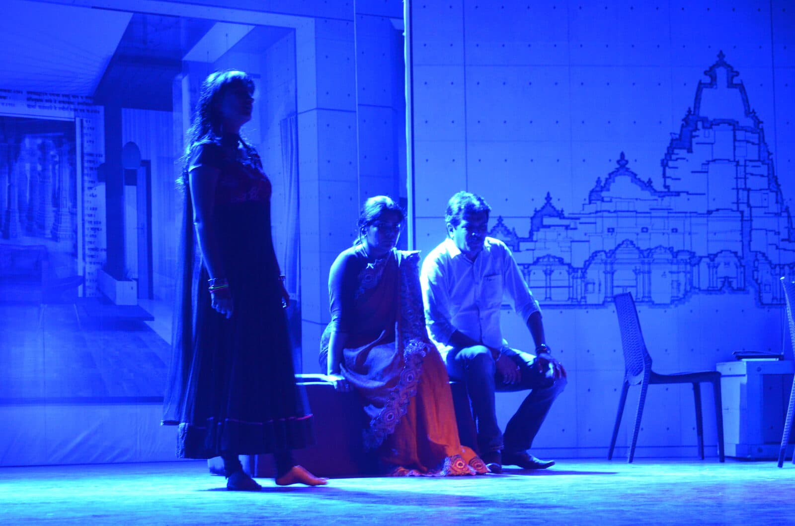 Three figures in deep blue light against the temple drawings of the studio wall